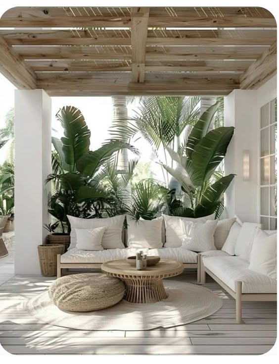An outdoor living room area under a light-colored wooden slatted pergola, with white sofa cushions, a round coffee table…