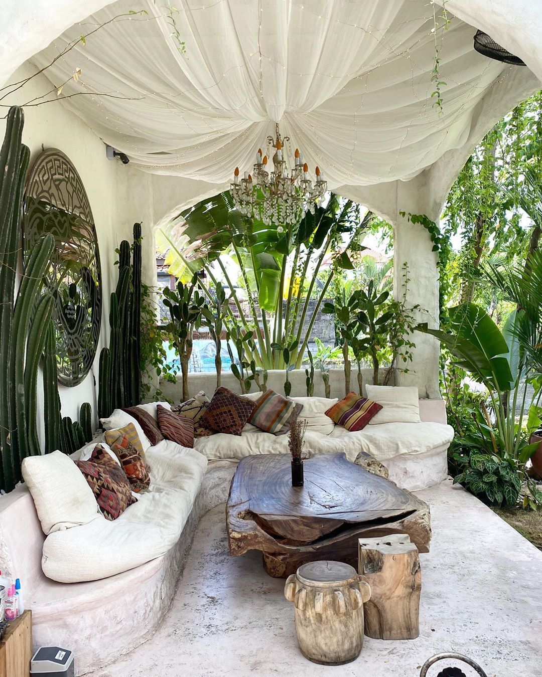 An outdoor living area with a curved white sofa, a large solid wooden coffee table, and an ornate chandelier draped with…