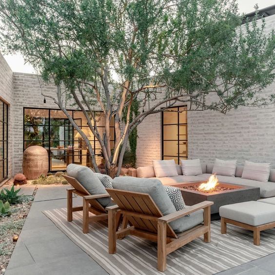 Outdoor patio with a large tree in the center, an L-shaped sofa around a square fire pit, and two wooden lounge chairs on a…