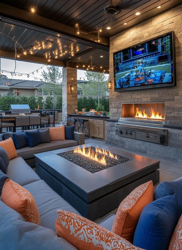 An outdoor living room with a large sectional sofa, a built-in fire pit, and a television mounted above an outdoor fireplace.