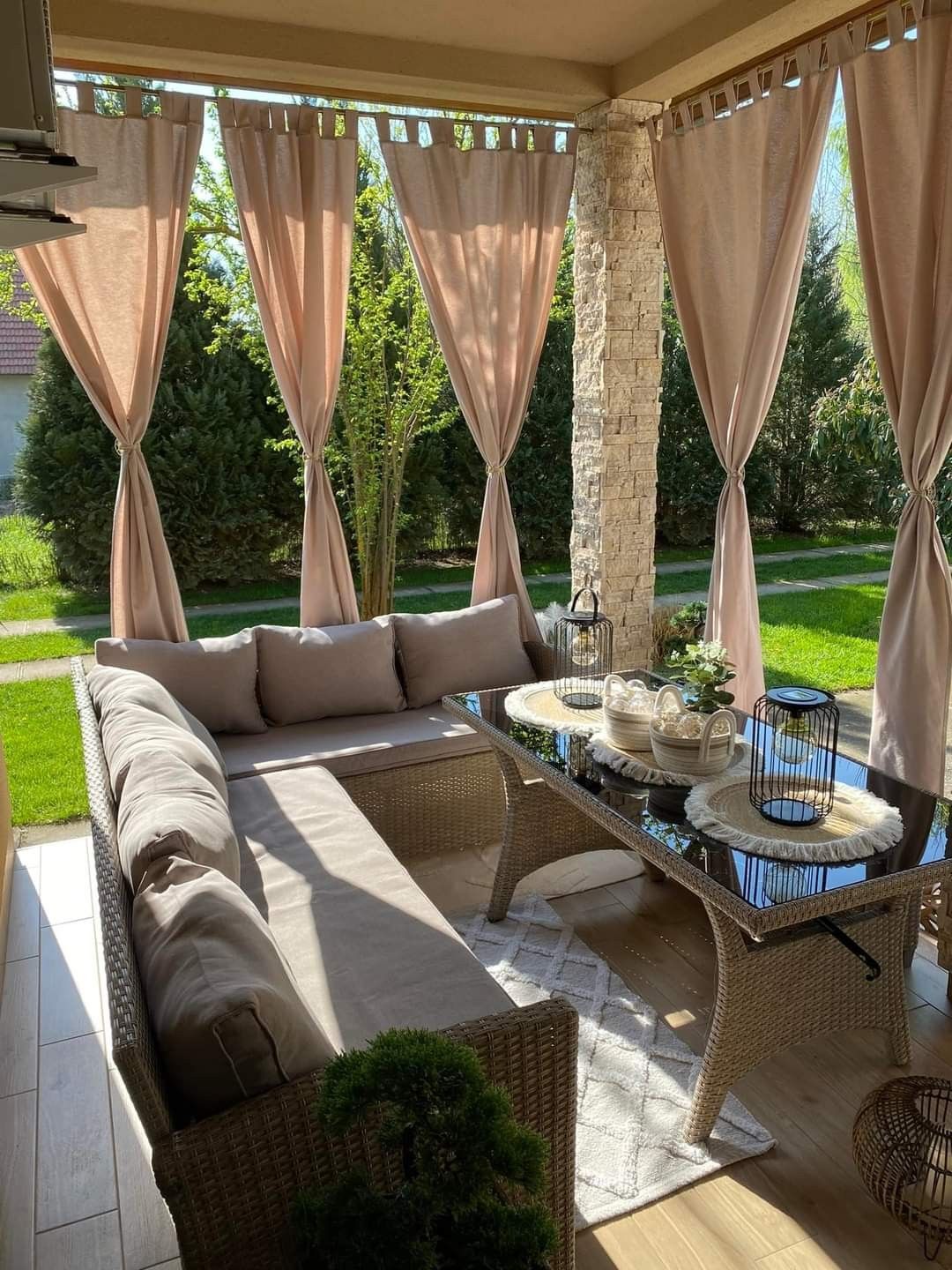 Covered outdoor patio with a large sectional sofa, glass-top table, and light-colored curtains.