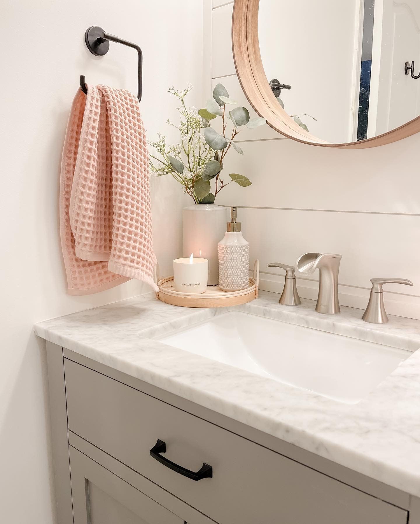 Bathroom vanity with marble counter, gray cabinet, white sink, and a round wooden mirror.