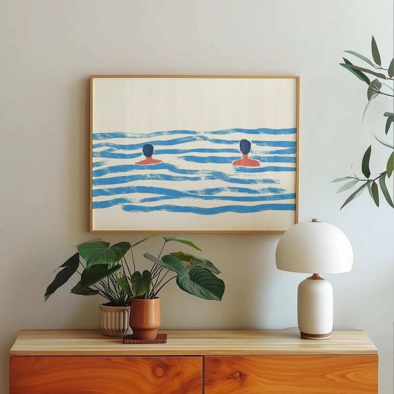 A framed art print of two people swimming in blue water hangs above a wooden cabinet with plants and a lamp.