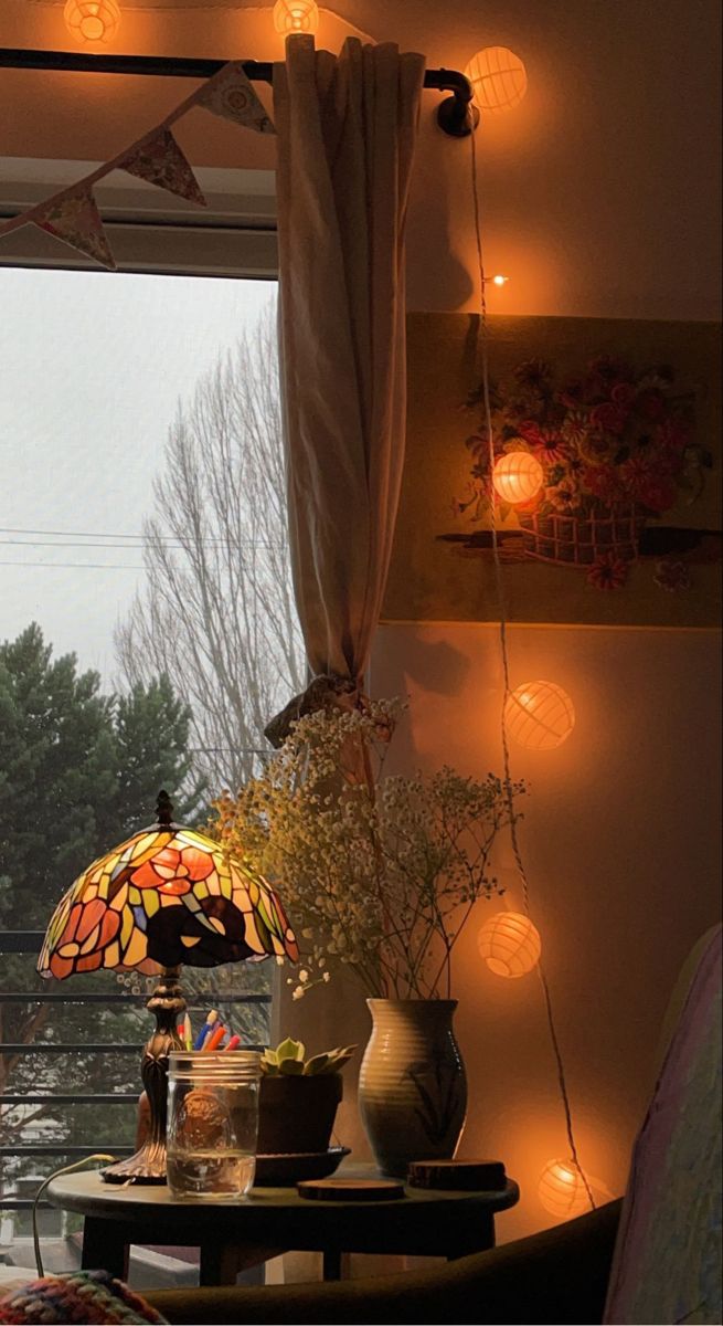 A stained-glass lamp and string lights illuminate a cozy interior with a window view of trees.