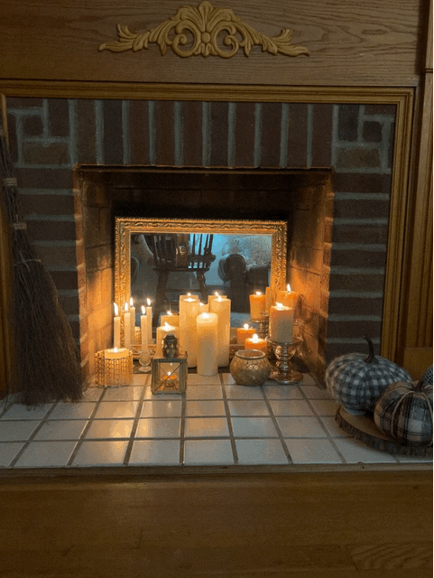 An assortment of lit candles and decorative lanterns arranged inside a fireplace opening, with a framed mirror behind them.
