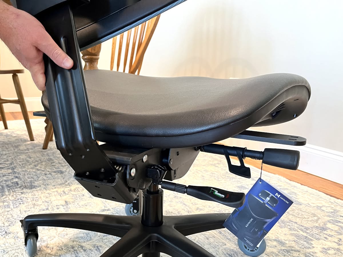 A person's hand adjusts a lever under the seat of a black ergonomic office chair, showing the seat pan mechanism.