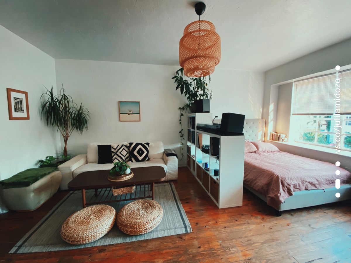 An open studio apartment featuring a low white bookshelf dividing the living room with a couch and coffee table from the…