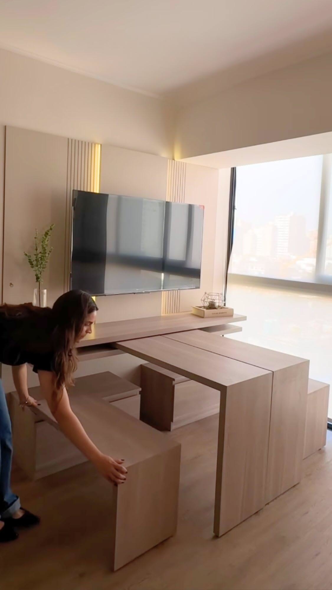 Person pulls out light wood benches and a table from a wall unit with a TV above, creating a dining area