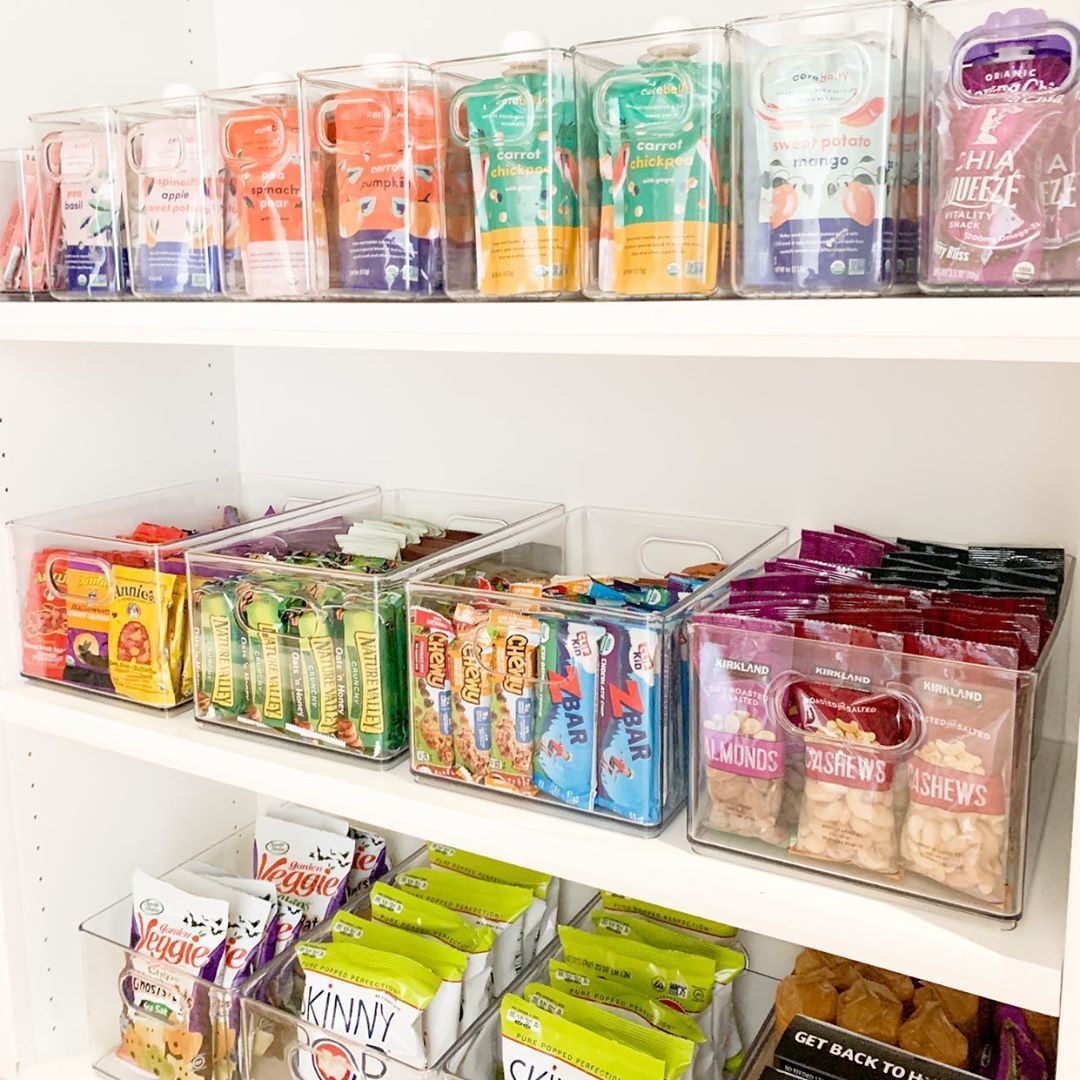Clear plastic bins hold various packaged snacks like fruit pouches, granola bars, and nut packs on white pantry shelves.