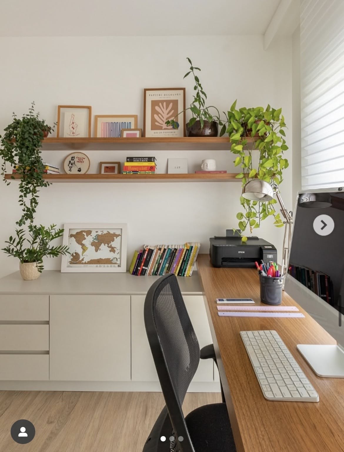 A home office features a wood desk and shelves, a black mesh chair, and several potted plants.