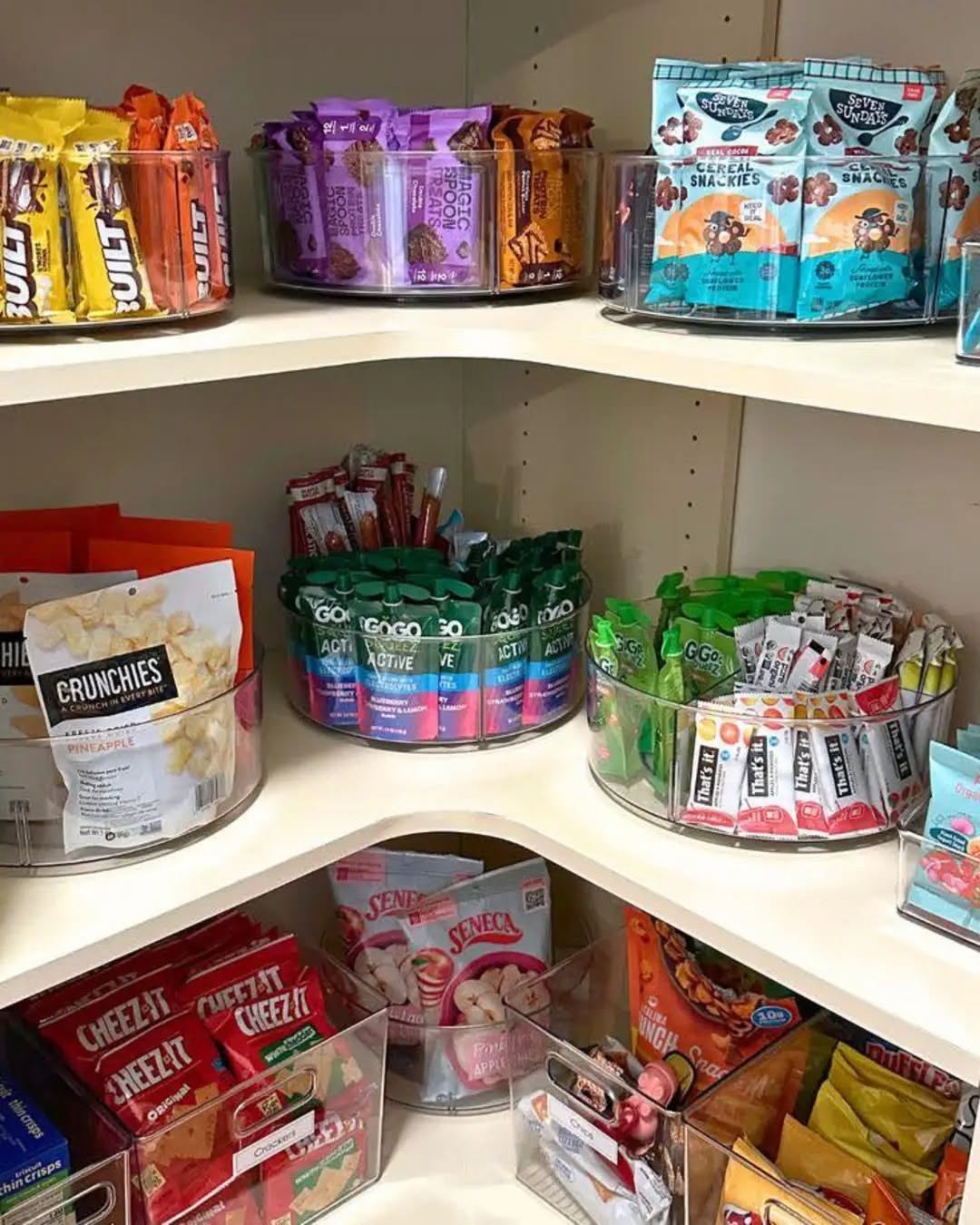 A pantry corner with multiple clear, segmented lazy Susan organizers filled with various packaged snacks like fruit pouches…