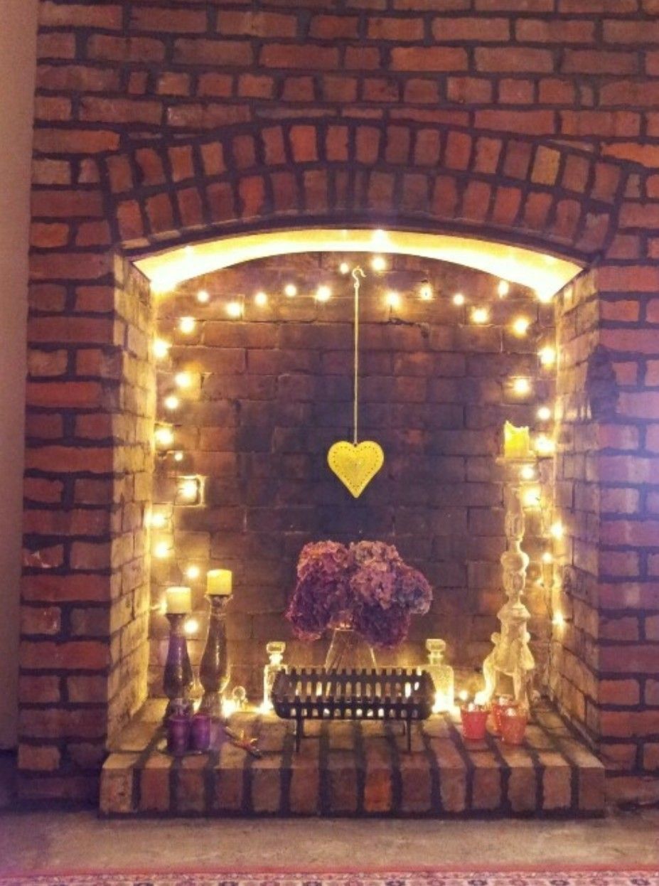 Non-working brick fireplace with string lights, candles, a floral arrangement, and a hanging heart decoration inside.