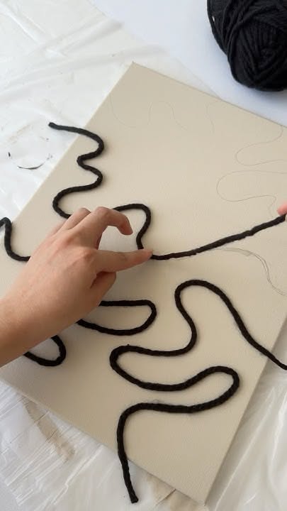 A person's hands arranging black yarn into a wavy pattern on a beige canvas board.