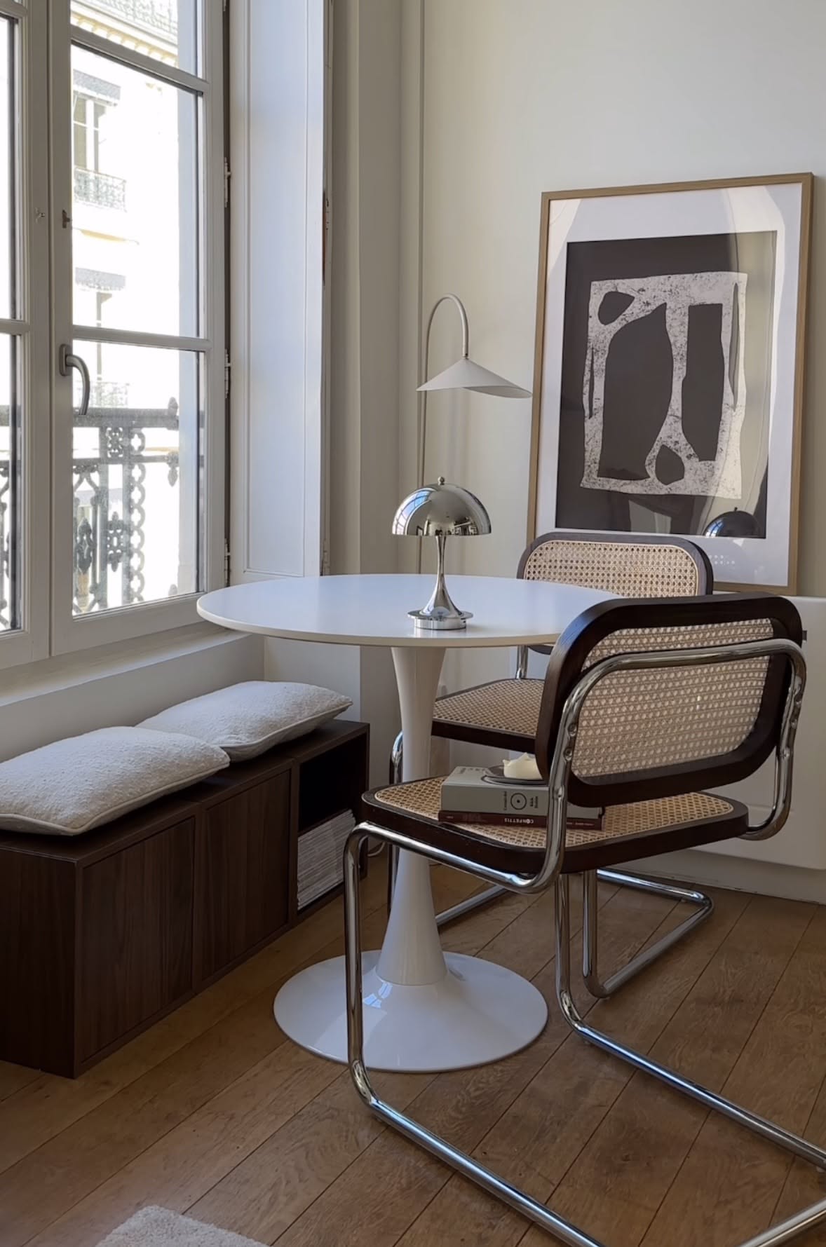 Small dining area with a round white table, two chairs, and a window nook bench with cushions.