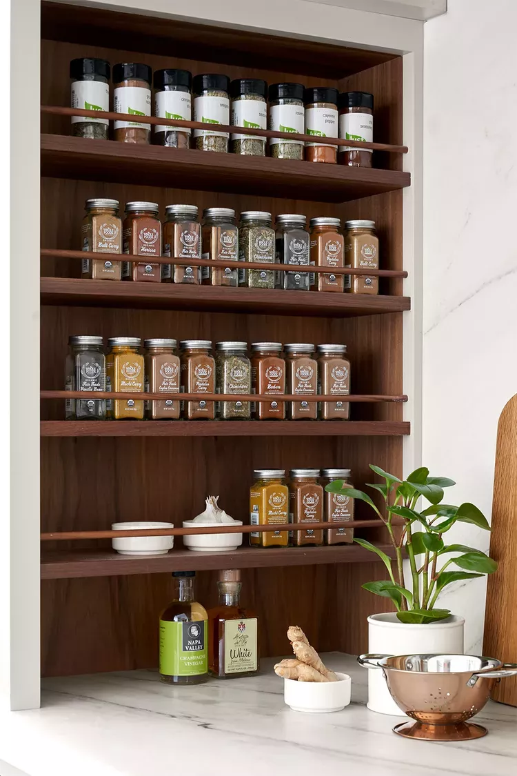 Built-in wooden spice rack with multiple shelves holding rows of small jars of spices, and a plant and vinegar bottles below.