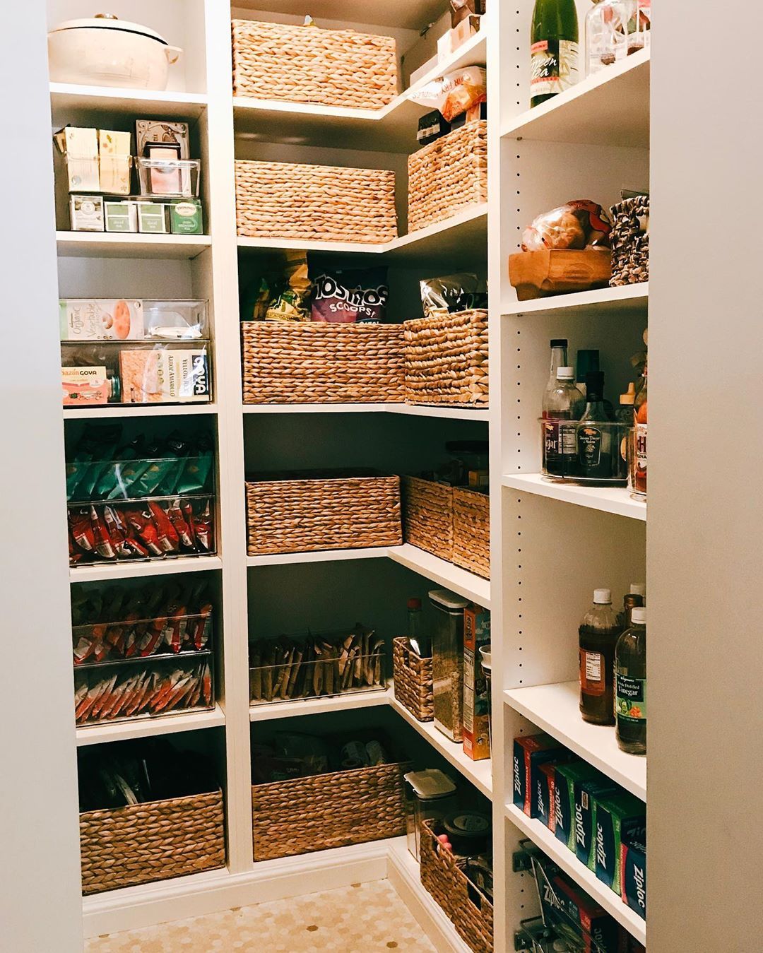 An organized pantry shows shelves filled with snack boxes, clear bins with snack bags, and woven baskets with various pantry…