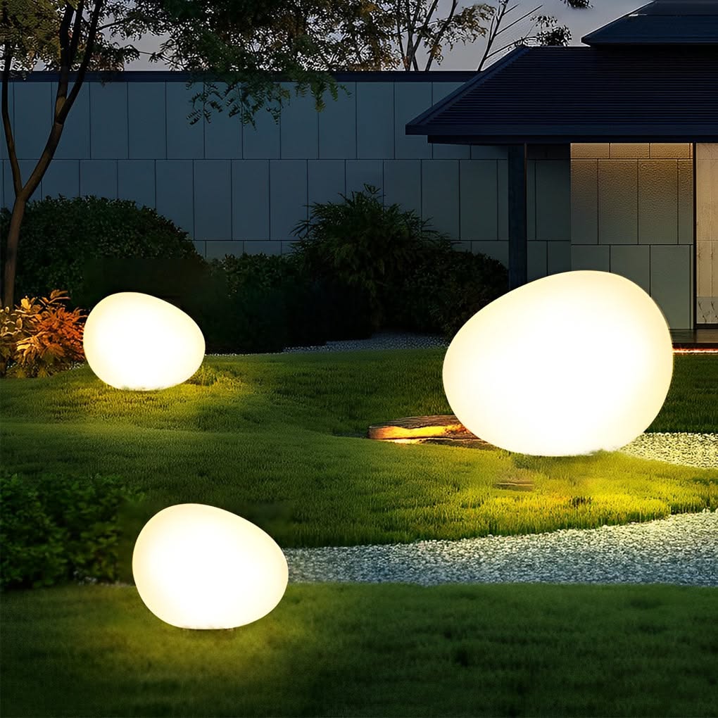 Three large, glowing white pebble lights nestled in green grass next to a gravel path at night.