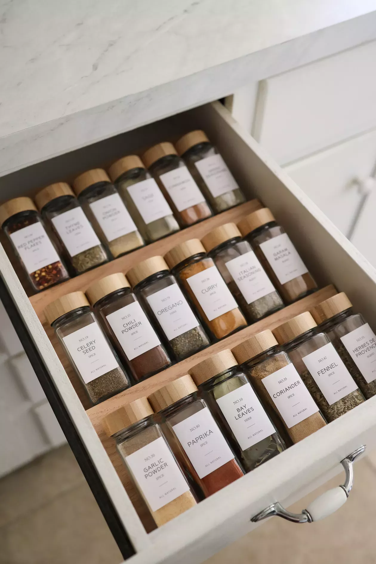 An open kitchen drawer containing multiple glass spice jars with wooden lids, arranged on a tiered wooden organizer.