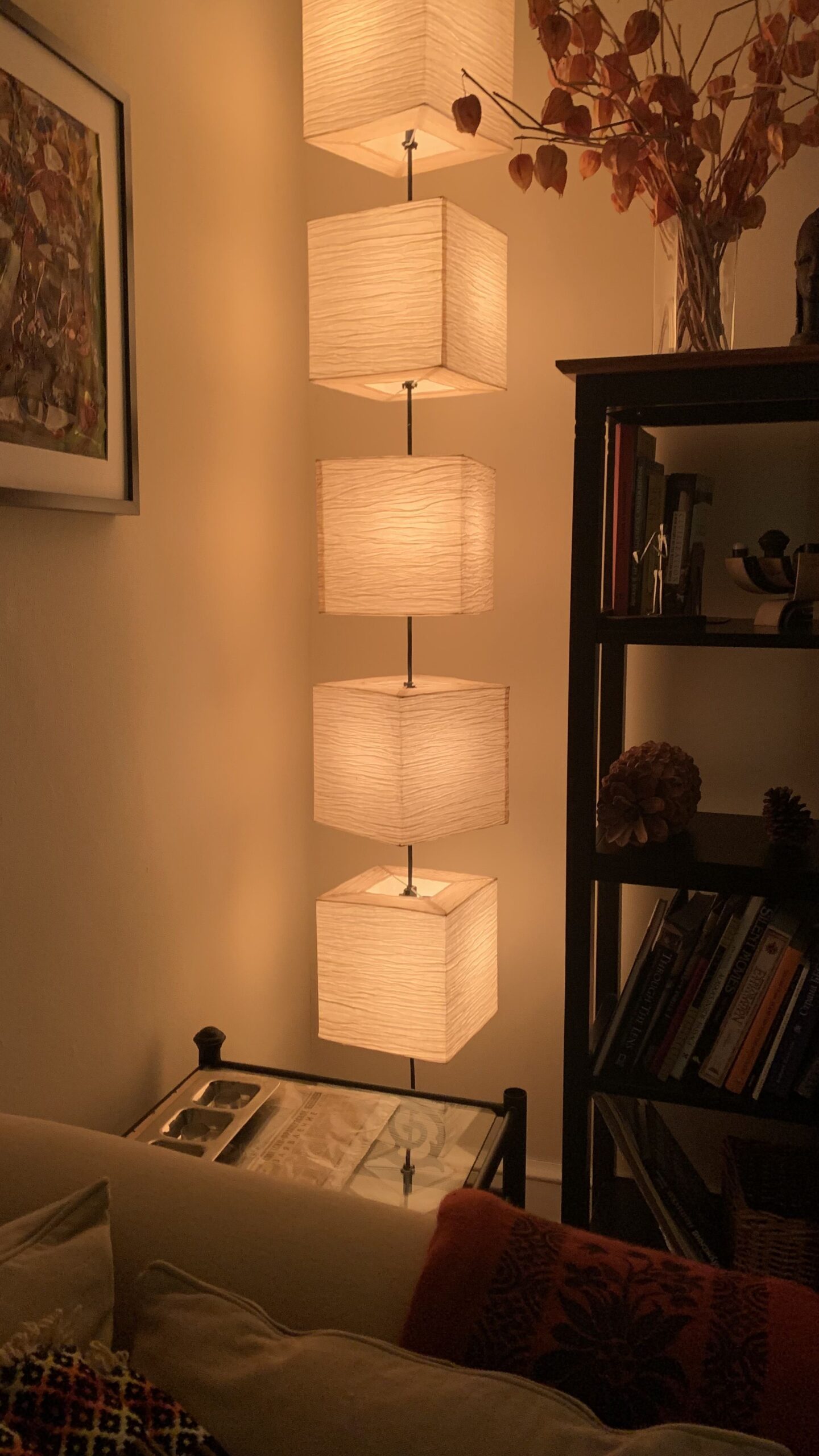 A floor lamp with five stacked, rectangular, fabric shades illuminates a cozy living room next to a bookcase and couch.