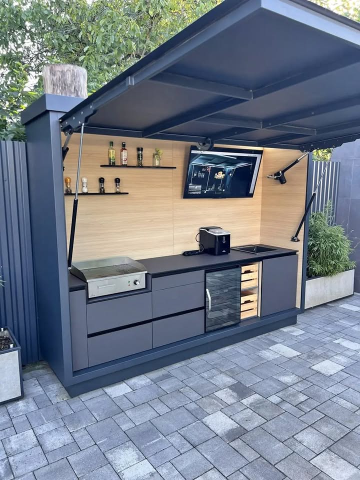 Compact outdoor kitchen unit with a flat-top grill, small sink, refrigerator, and storage drawers under a protective canopy.