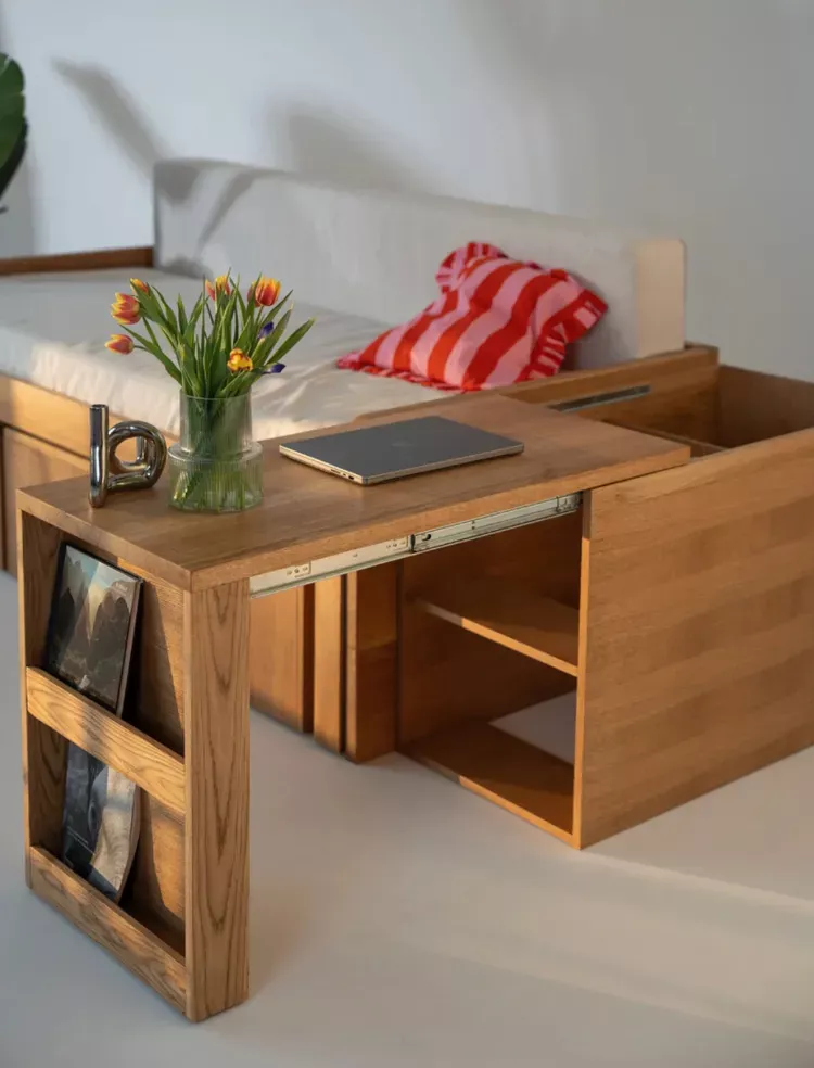 Wooden bed with a mattress and a sliding wooden desk pulled out, featuring shelves and a laptop on top.