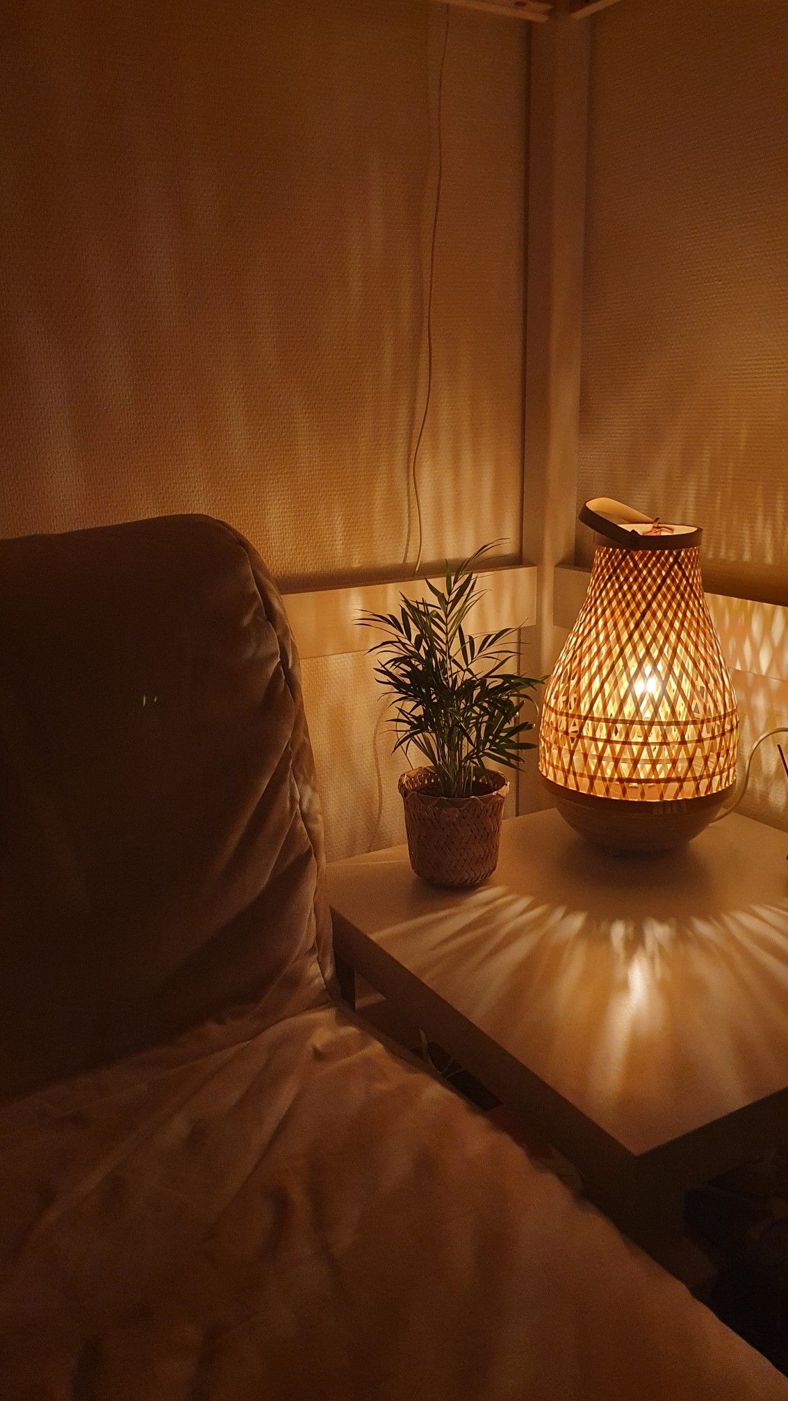 A bedside table with a woven bamboo lamp casting patterned shadows on the wall and table, next to a small potted plant and a…