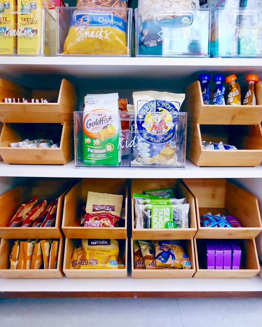 A pantry shelf with multiple wooden stackable bins, each containing various individually packaged snack items like cookies…