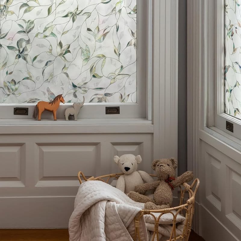 Close-up of a window covered with a translucent film featuring a leafy green pattern, next to a wicker basket with stuffed…