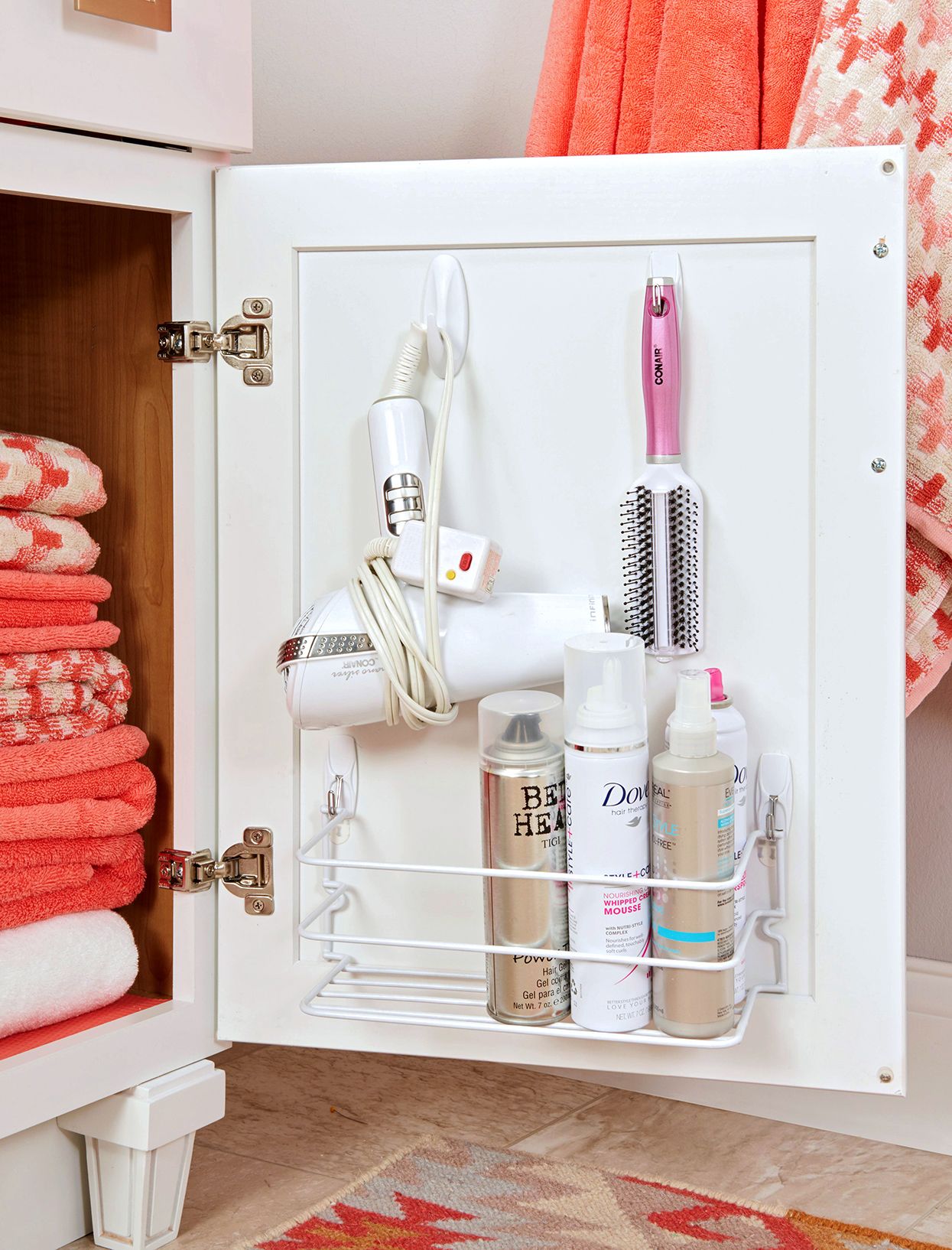 Open white bathroom cabinet door with a hair dryer, curling brush, and hair product bottles organized in a hanging wire…