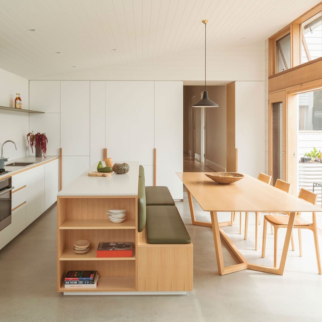 Modern kitchen island with open shelving and an attached green upholstered bench seating, facing a wooden dining table.