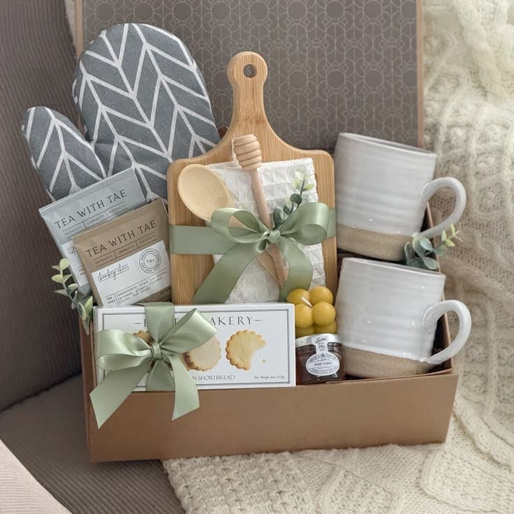 Gift basket with gray patterned oven mitts, small wooden cutting board, two mugs, tea, shortbread cookies, and a honey…