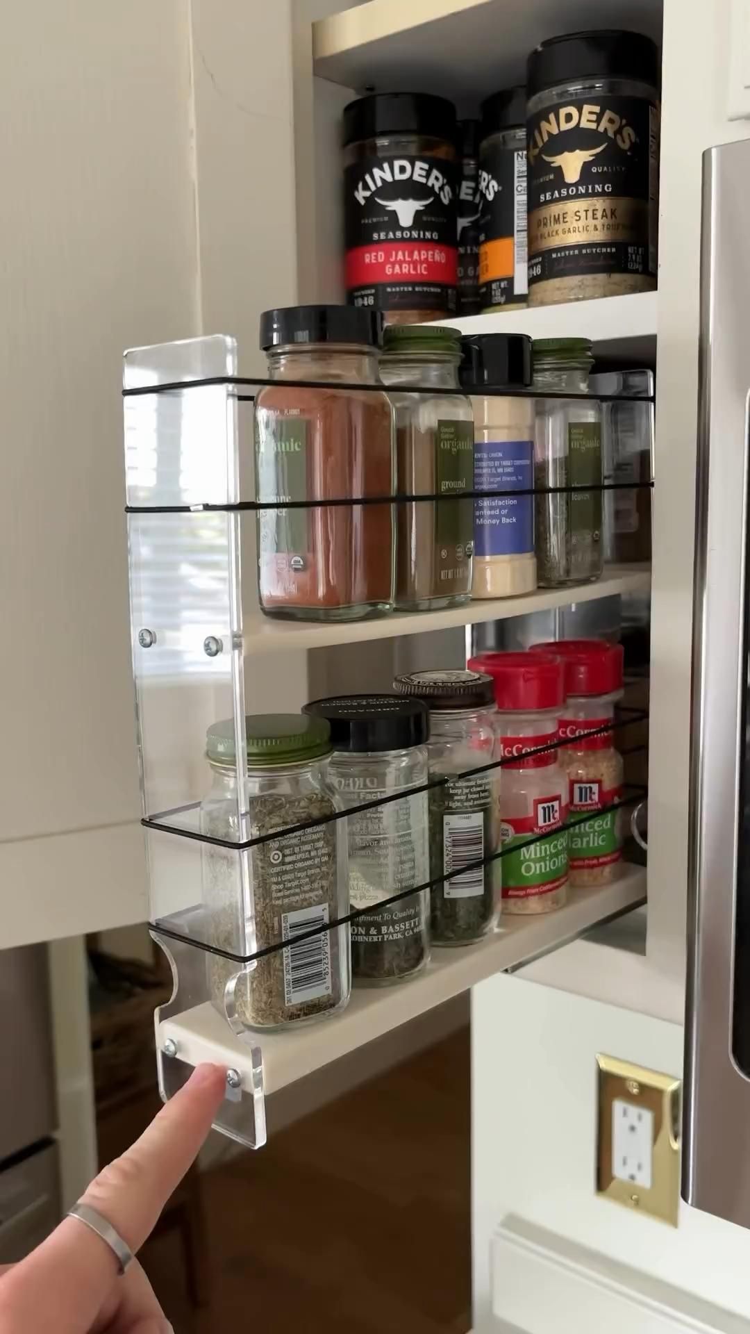 A person pulls out a clear, multi-tiered spice organizer, filled with assorted spice jars, from inside a kitchen cabinet.