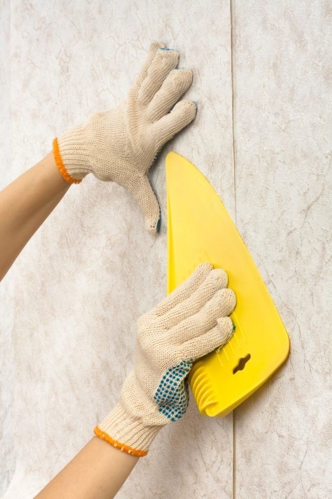 Hands in work gloves using a yellow smoothing tool on newly applied wallpaper