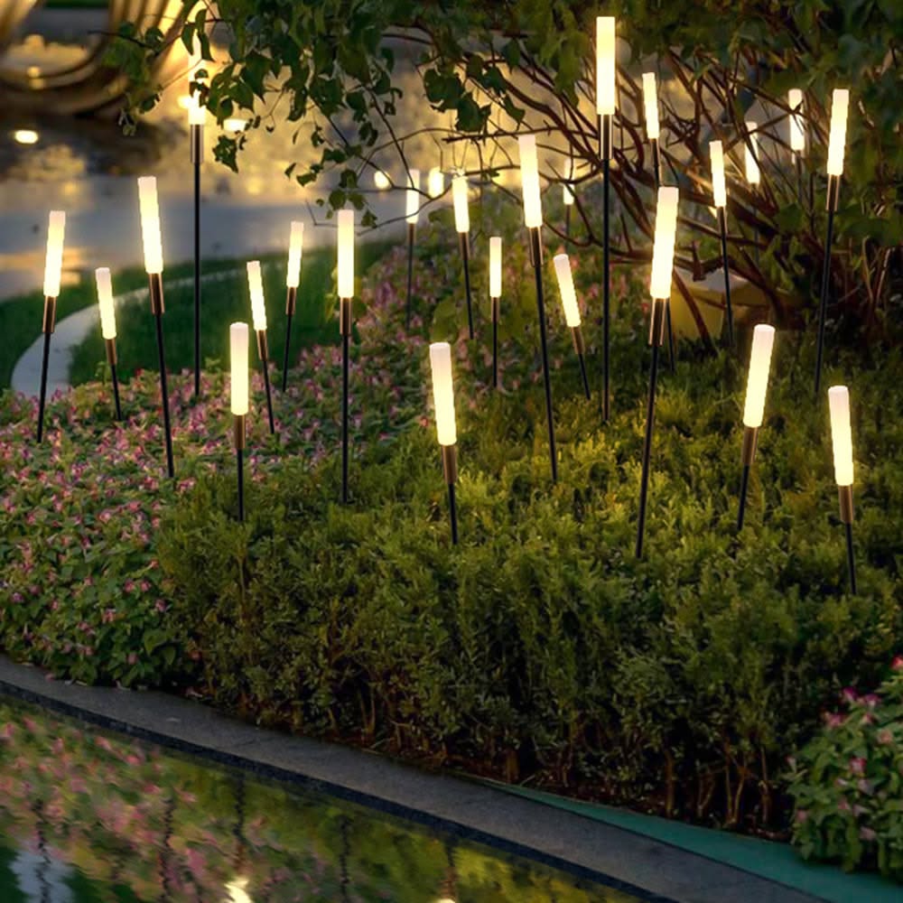 Multiple modern solar spike lights with glowing tops arranged among green garden bushes and small pink flowers, next to a…
