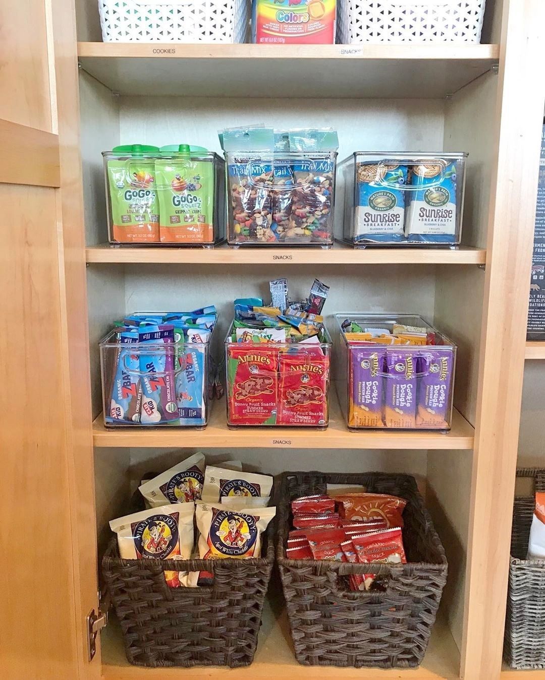 Pantry shelves organized with clear bins and woven baskets full of various packaged snacks like fruit pouches, trail mix…