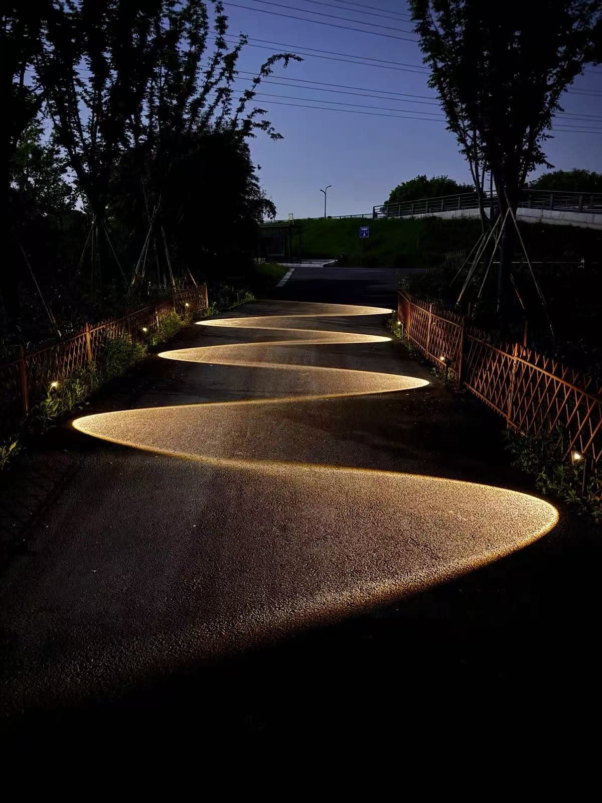 A dark outdoor path at dusk illuminated by a wavy, S-shaped pattern of warm light from ground-level fixtures.