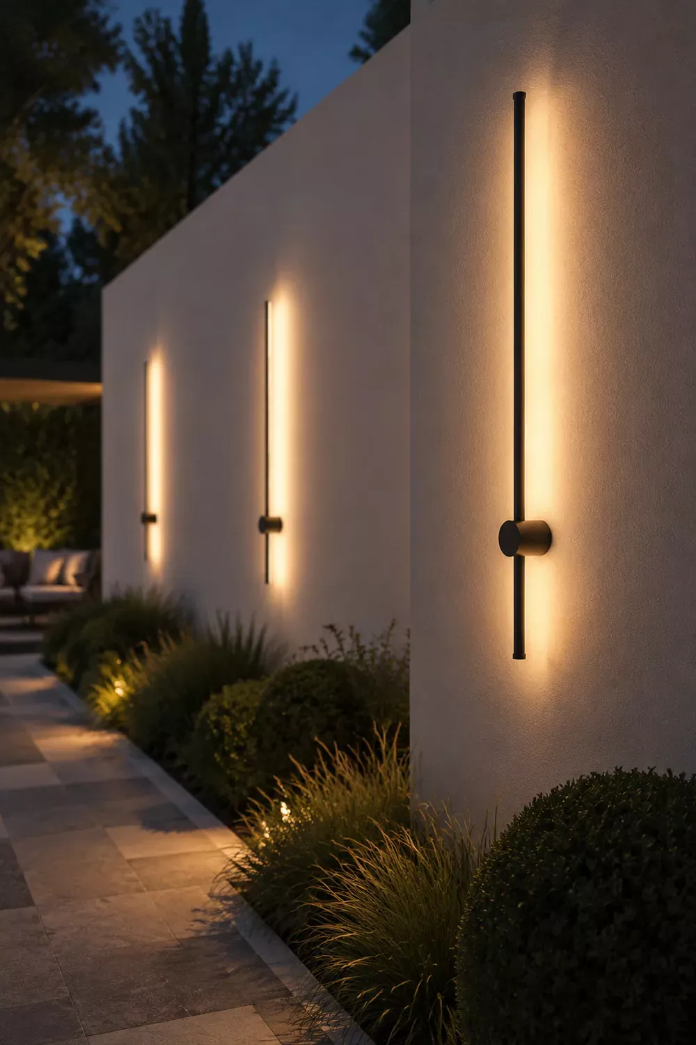 Three vertical linear wall lights illuminate a modern outdoor wall and adjacent landscaping at dusk.