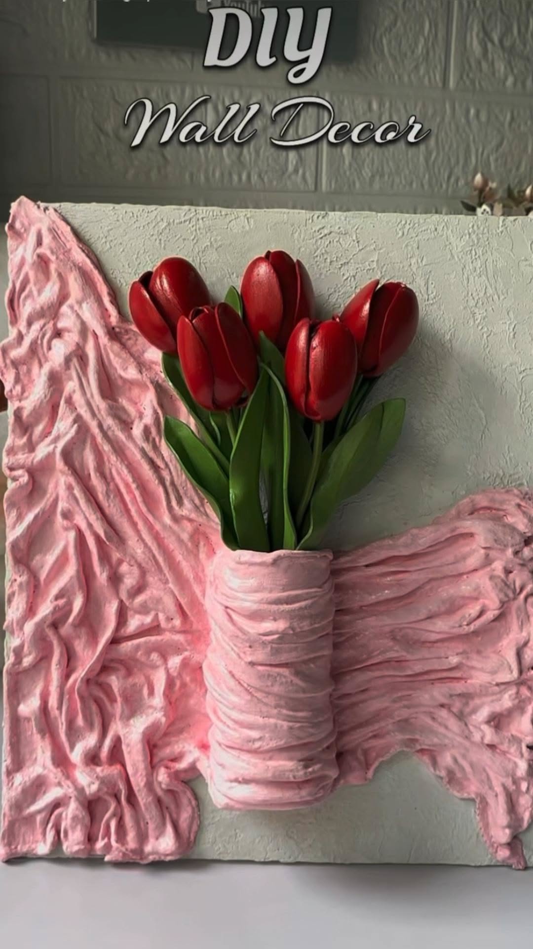 Decorative wall art featuring textured pink fabric or plaster arranged as a vase, holding a bouquet of red tulips against a…