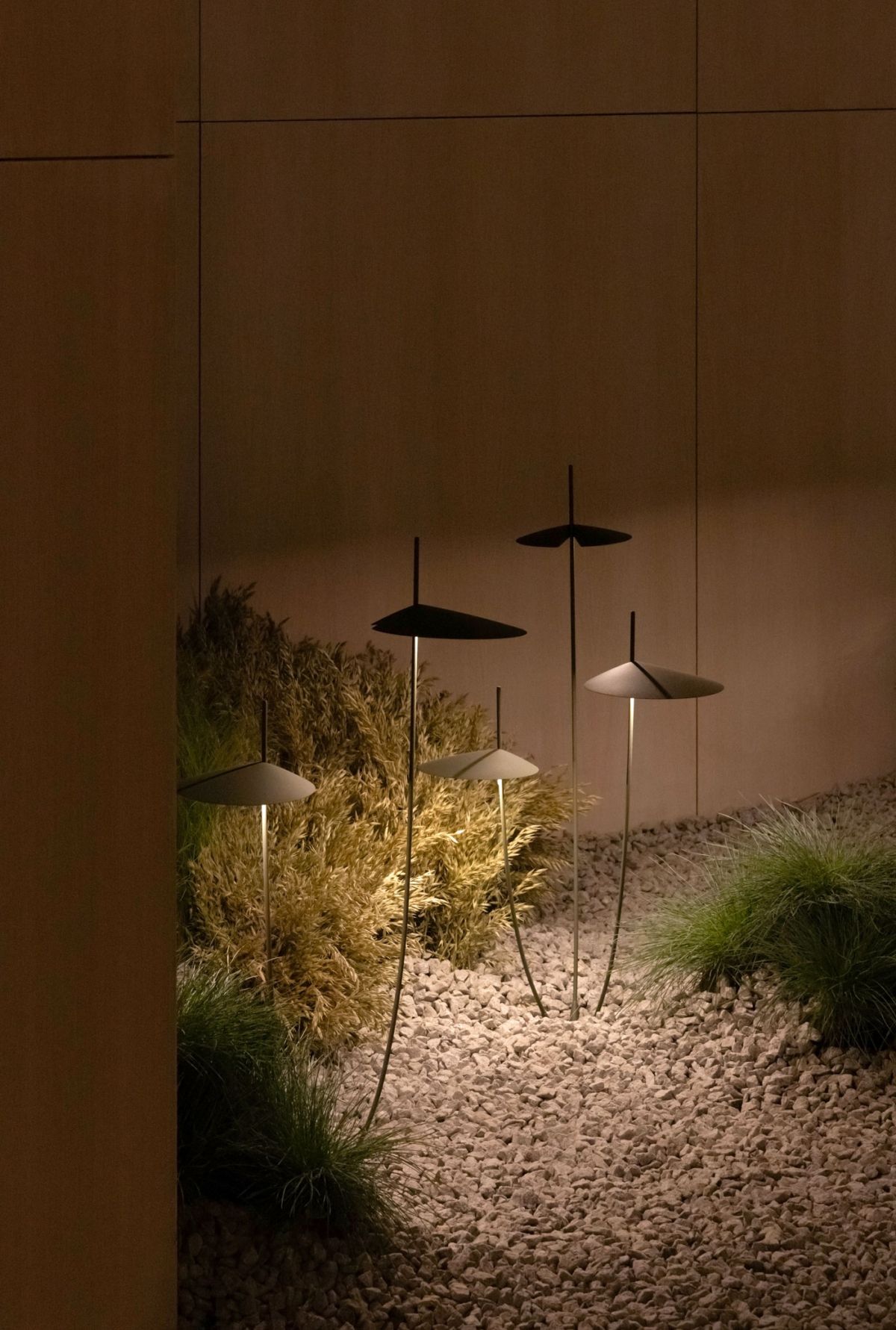Several modern, dark-colored mushroom-shaped ground lights with thin stems among ornamental grasses and pebbles at night.