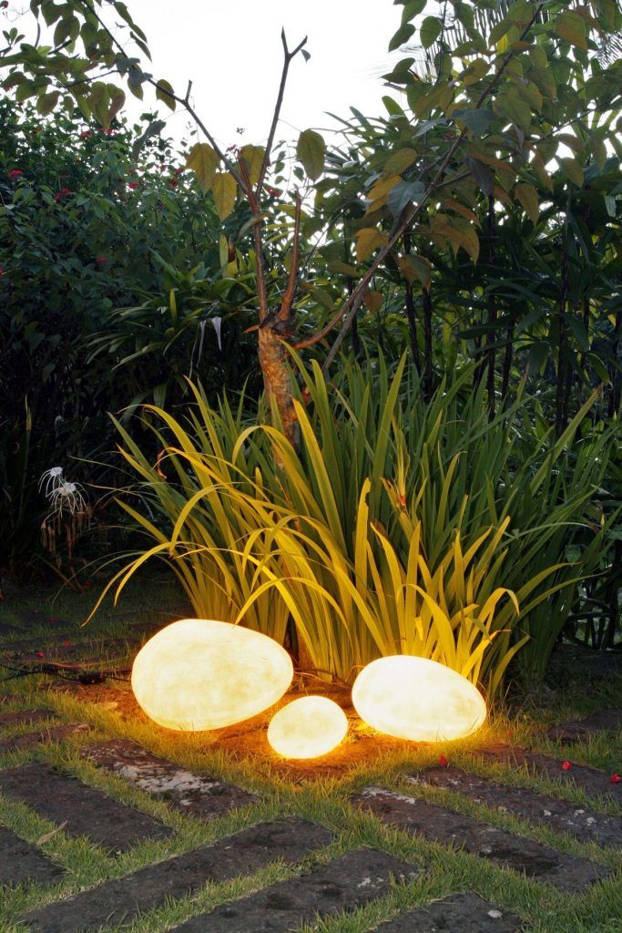 Three illuminated, pebble-shaped lights glowing warmly amidst tall green foliage and a stone path.