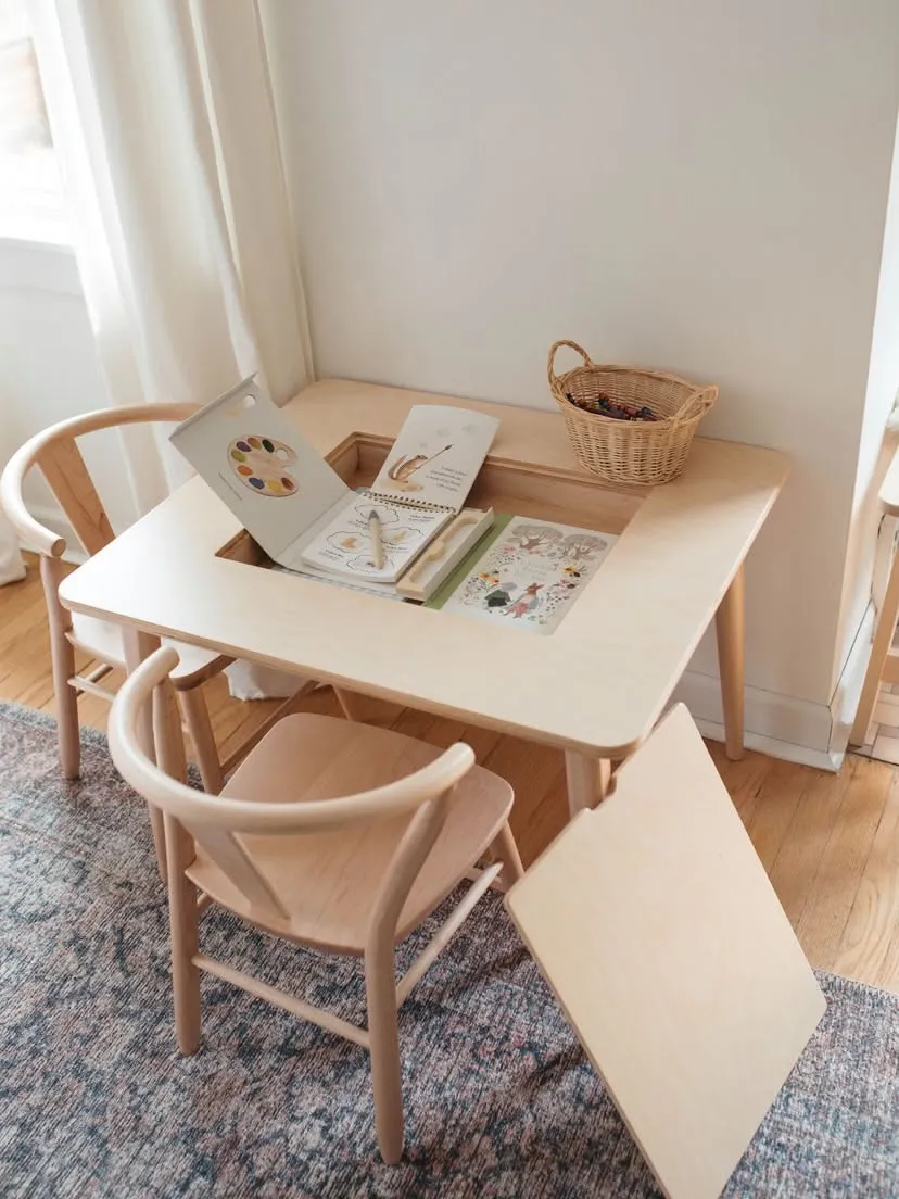 Light wood children's table with a central compartment holding art supplies, two matching chairs, and a small basket.