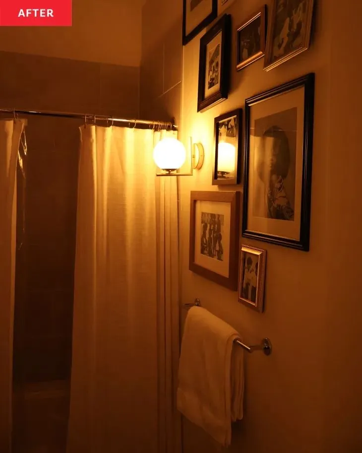 A round, white glass wall sconce illuminates a collection of framed artwork on a bathroom wall next to a shower curtain.