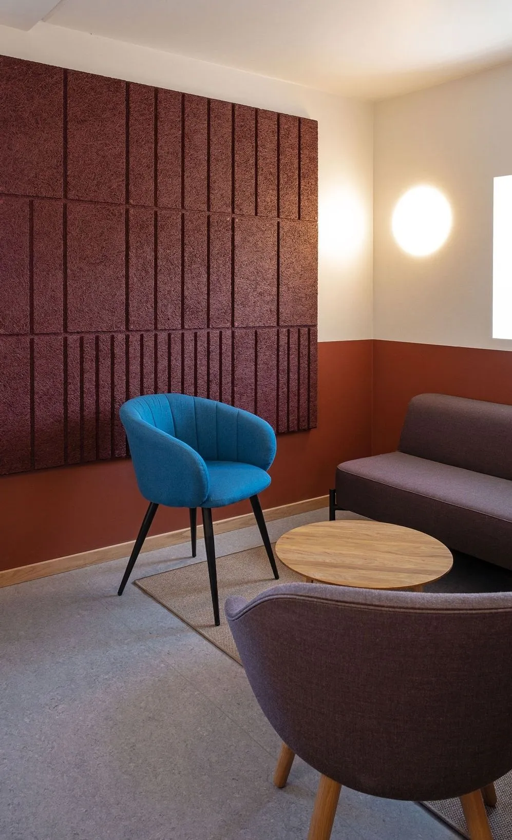 Lounge area with a large, textured burgundy acoustic panel installation on one wall, a blue armchair, and a grey sofa.