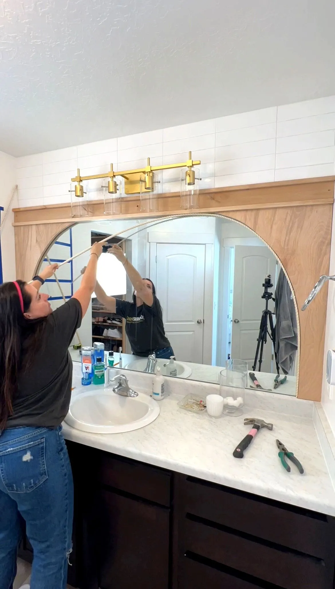 Woman applying white rope trim along the curved edge of a large bathroom mirror mounted above a vanity.