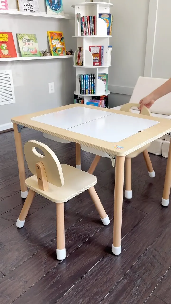 Wooden children's table with two white-topped panels that open, accompanied by two small wooden chairs, in a bright room.