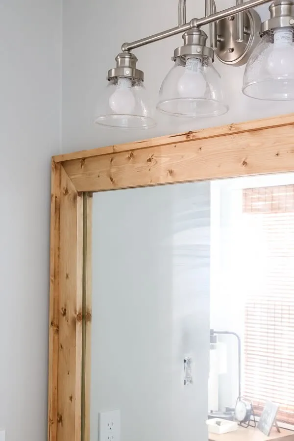 Close up of a builder-grade bathroom mirror framed with light-colored, knotty wood trim, under a three-light vanity fixture.