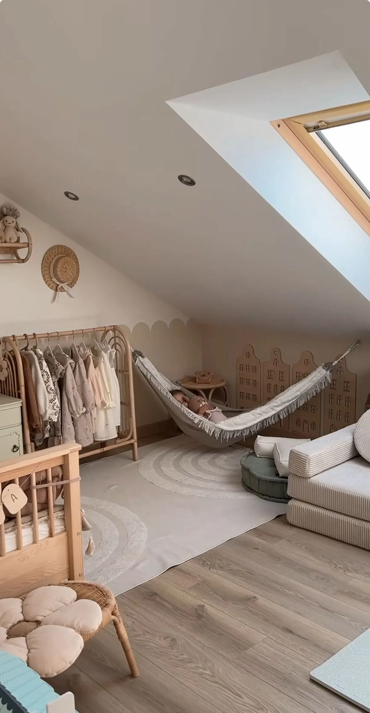 Nursery room with sloped ceiling, crib, clothes rack, child in a hammock, and floor seating on a light rug.