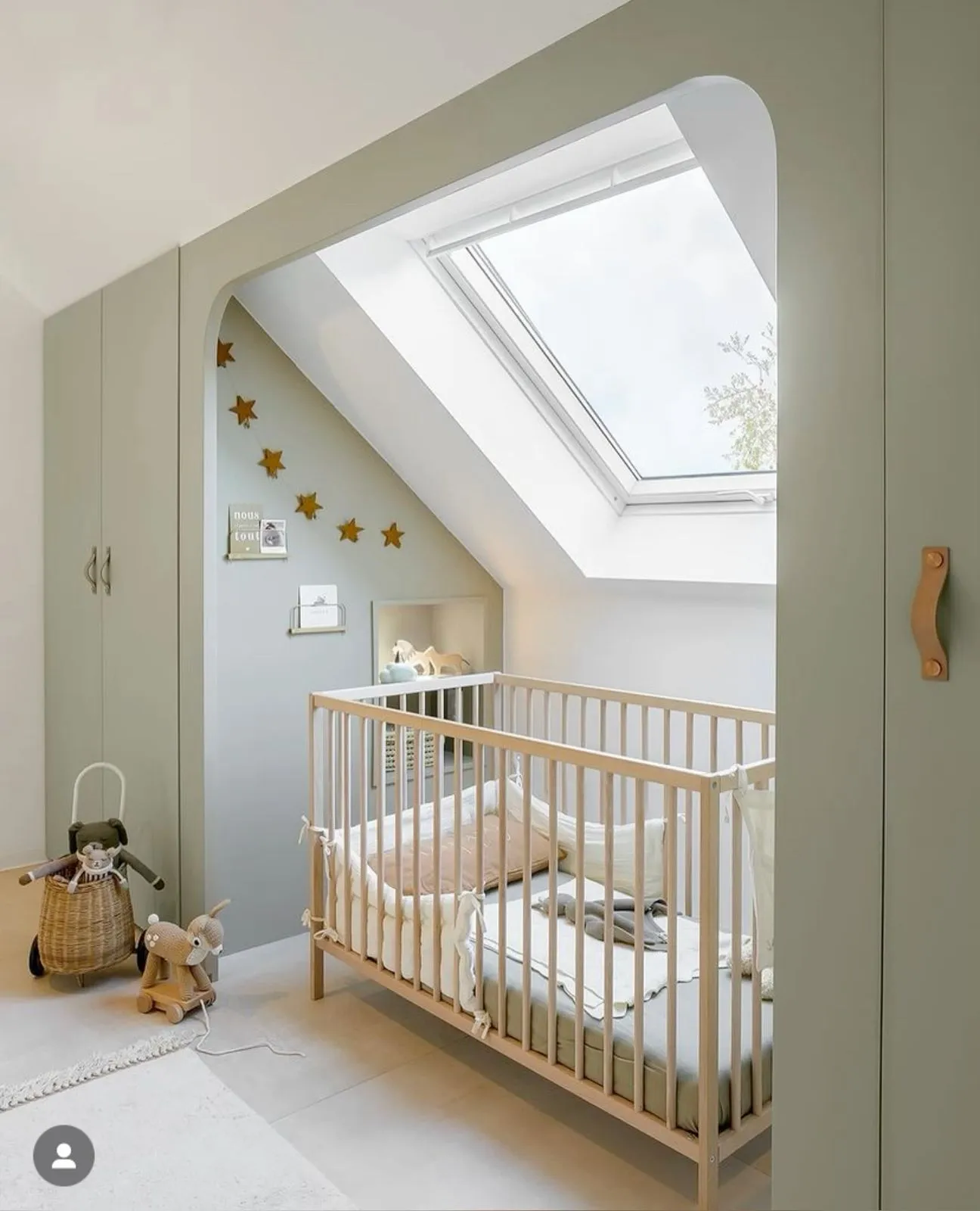 Baby crib in an attic nursery with custom sage green built-in wardrobes and an arched window opening above the crib.