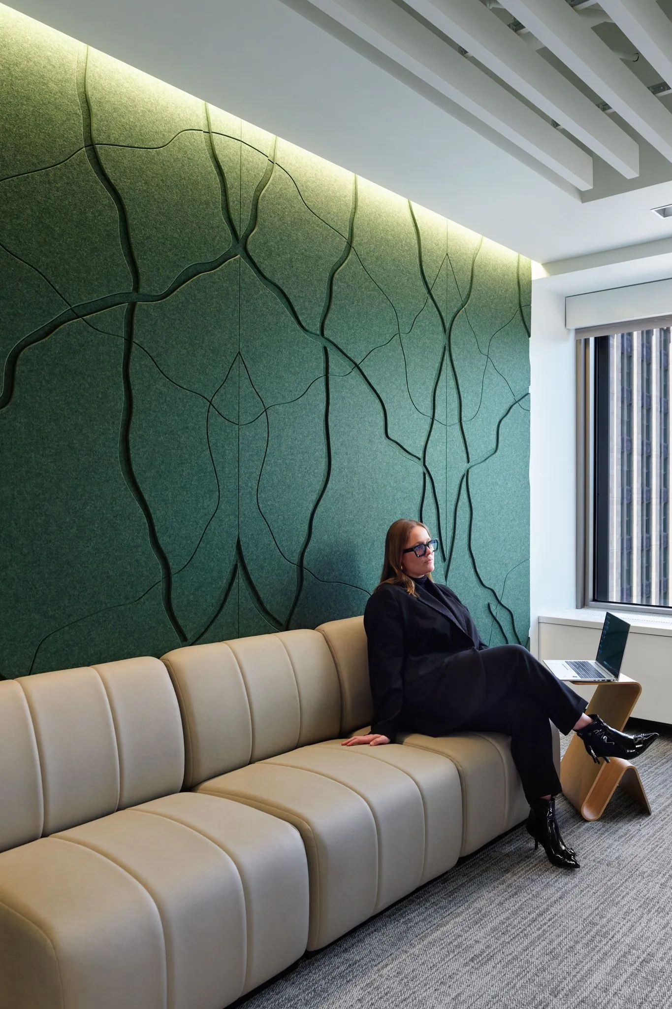 Woman in black suit sitting on a light-colored sofa in front of a green textured acoustic wall panel.