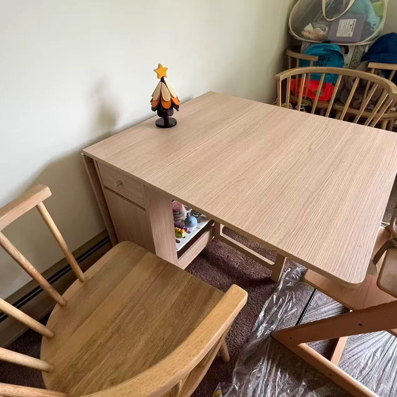 Light wood drop-leaf table with one leaf extended, showing a small drawer and shelves underneath. A wooden chair is nearby.